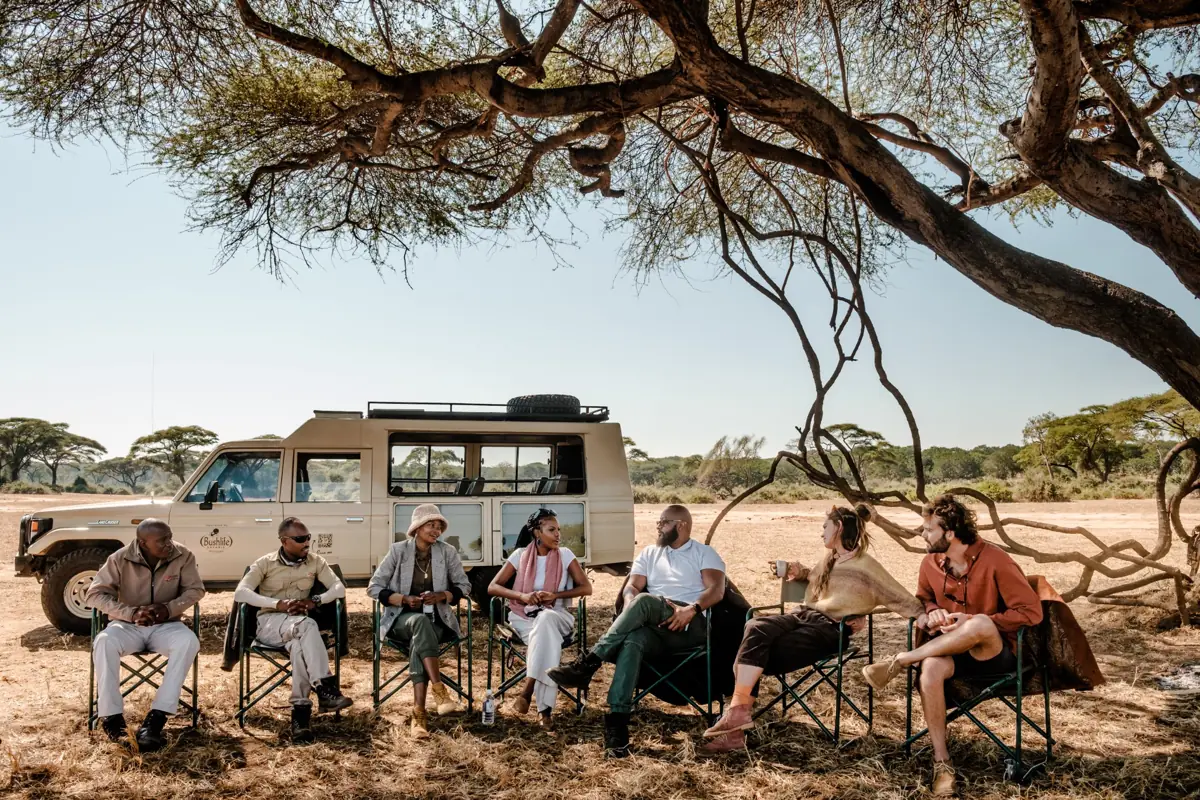 Guests and guides relaxing under a tree beside a safari vehicle