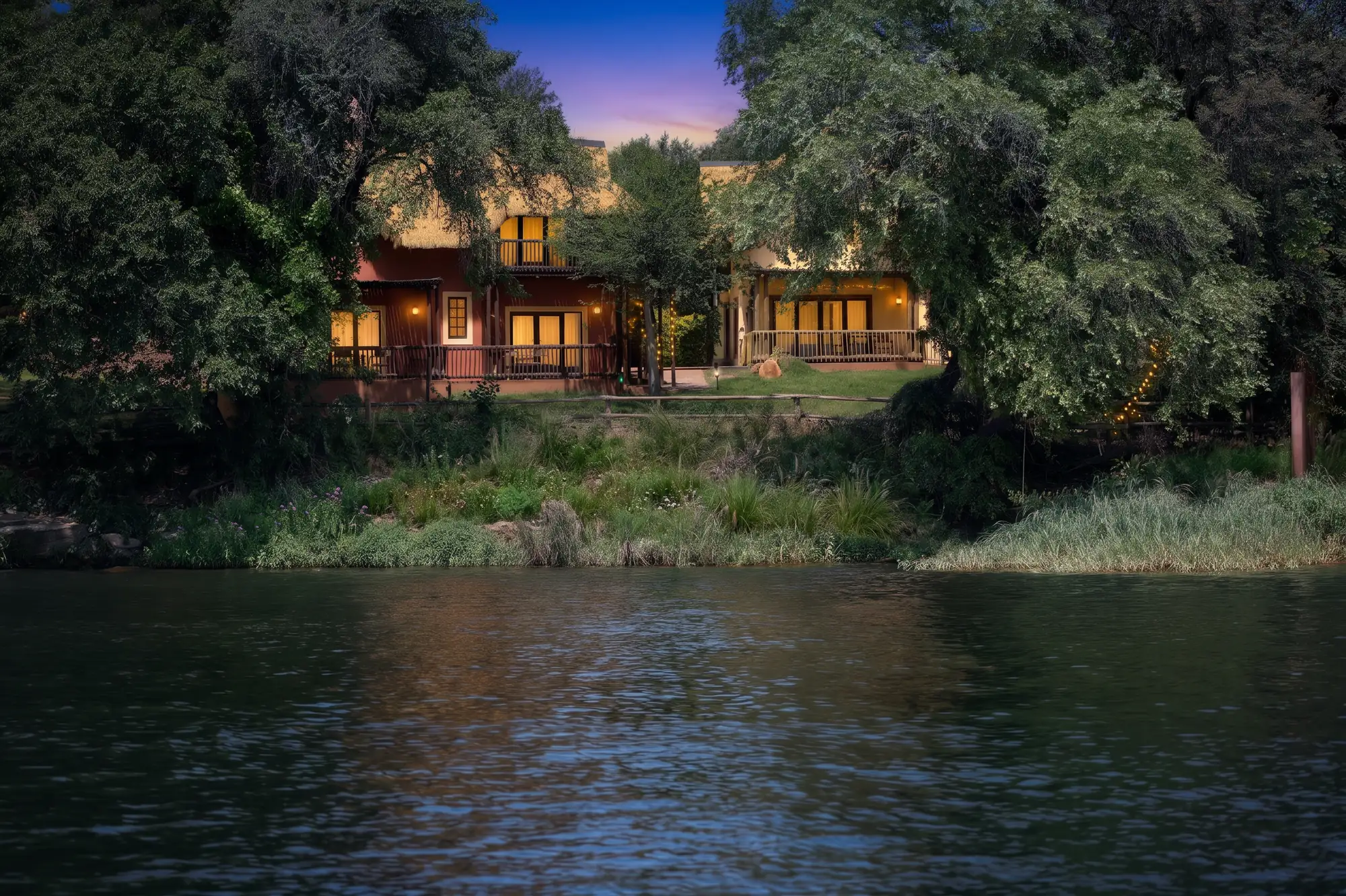 Thatched chalets glowing at dusk among the trees on the banks of the Chobe River, seen from the water