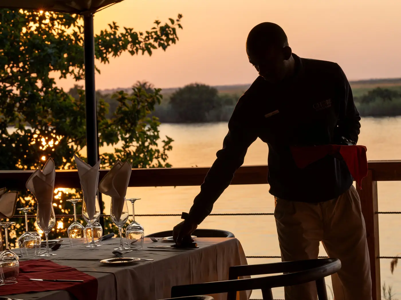 A waiter sets a table on the river deck as the sun sets over the Chobe